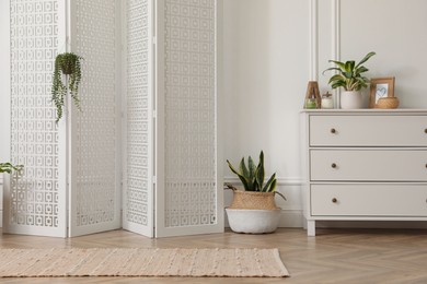 Stylish room with wooden folding screen and chest of drawers near white wall. Interior design