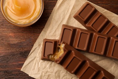 Tasty chocolate bars and bowl of caramel on wooden table, flat lay