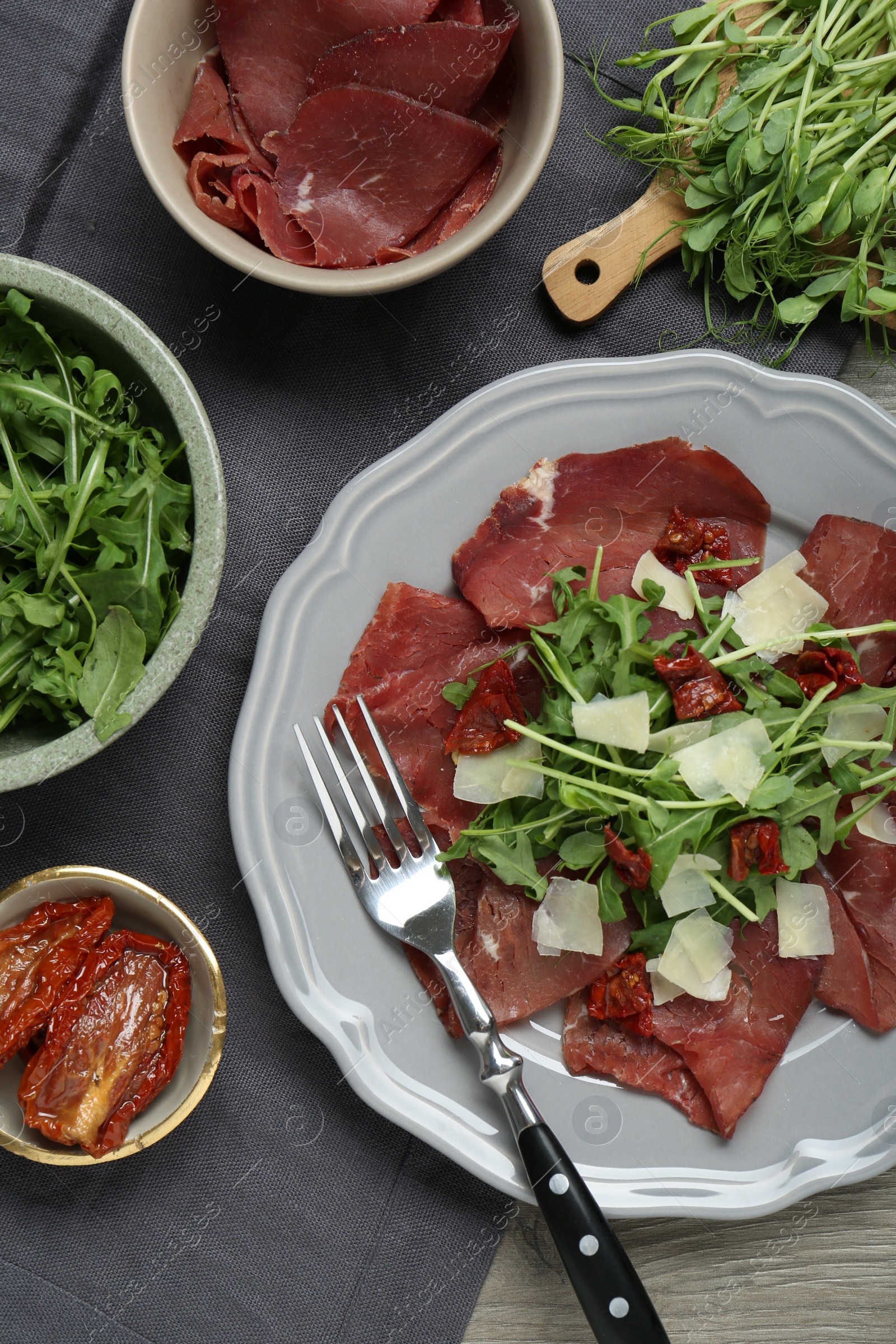 Photo of Plate of tasty bresaola salad with sun-dried tomatoes, parmesan cheese and cutlery on wooden table, flat lay