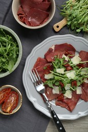 Photo of Plate of tasty bresaola salad with sun-dried tomatoes, parmesan cheese and cutlery on wooden table, flat lay