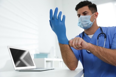 Photo of Doctor in protective mask putting on medical gloves at table in office