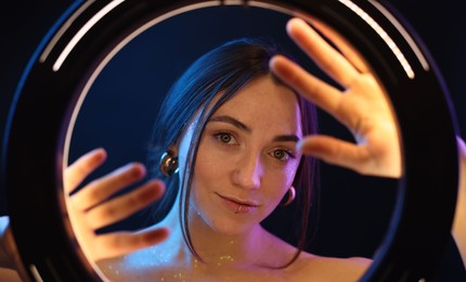 Photo of Portrait of beautiful woman on dark background in neon lights, view through ring lamp