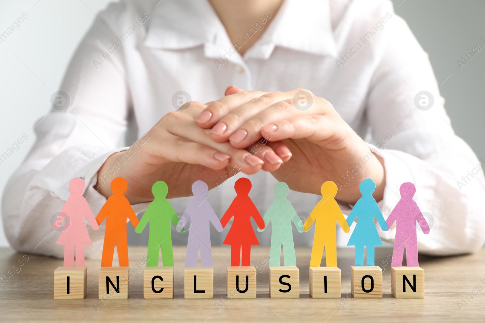 Photo of Woman protecting paper human figures and wooden cubes with word Inclusion at table, closeup