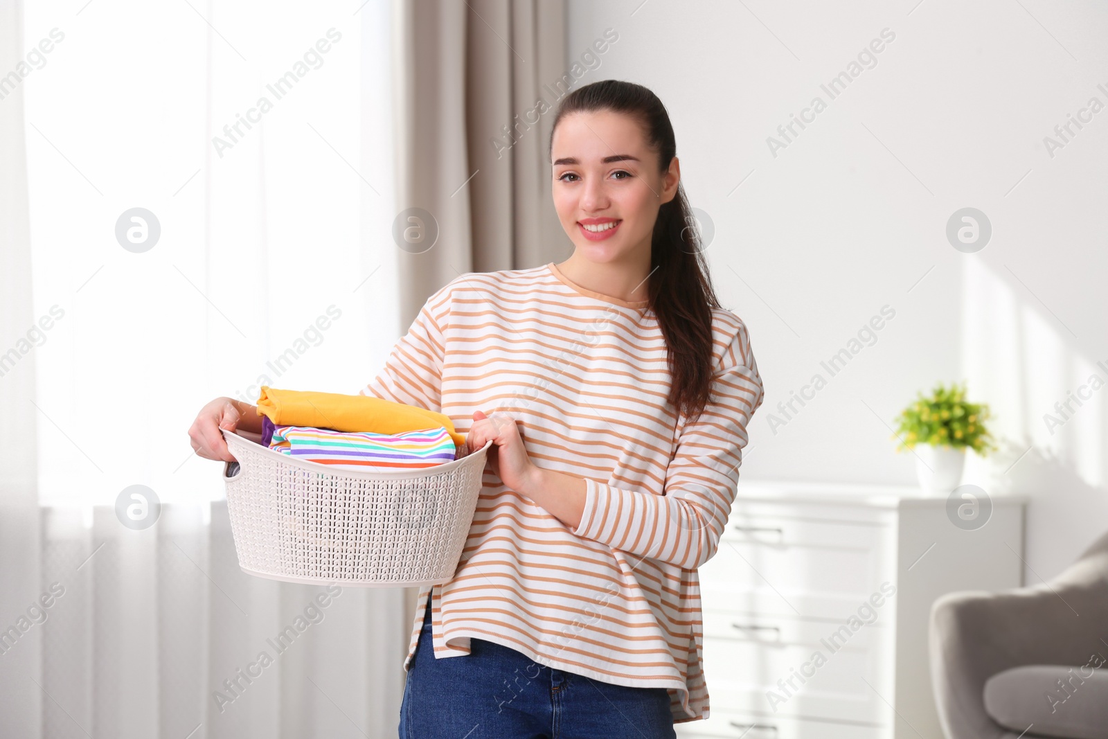 Photo of Happy young woman holding basket with laundry at home