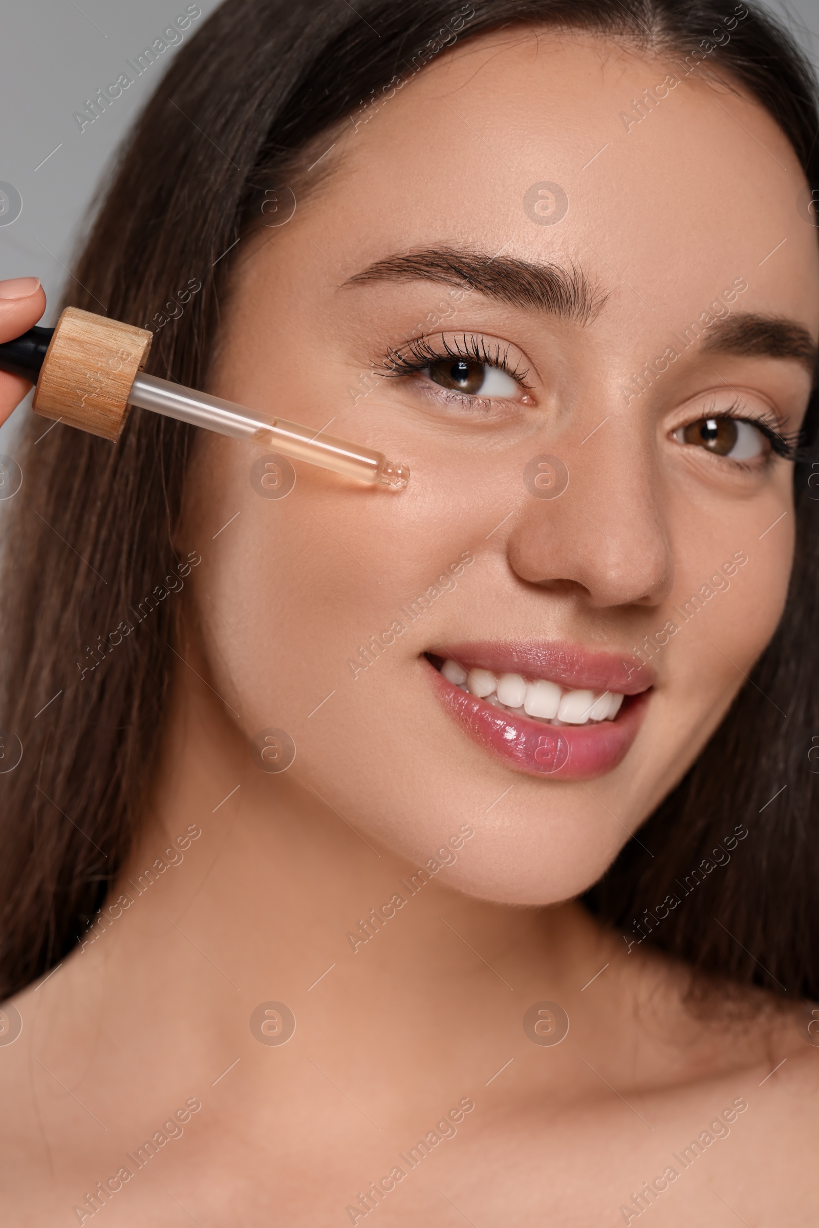 Photo of Beautiful young woman applying essential oil onto face on grey background, closeup