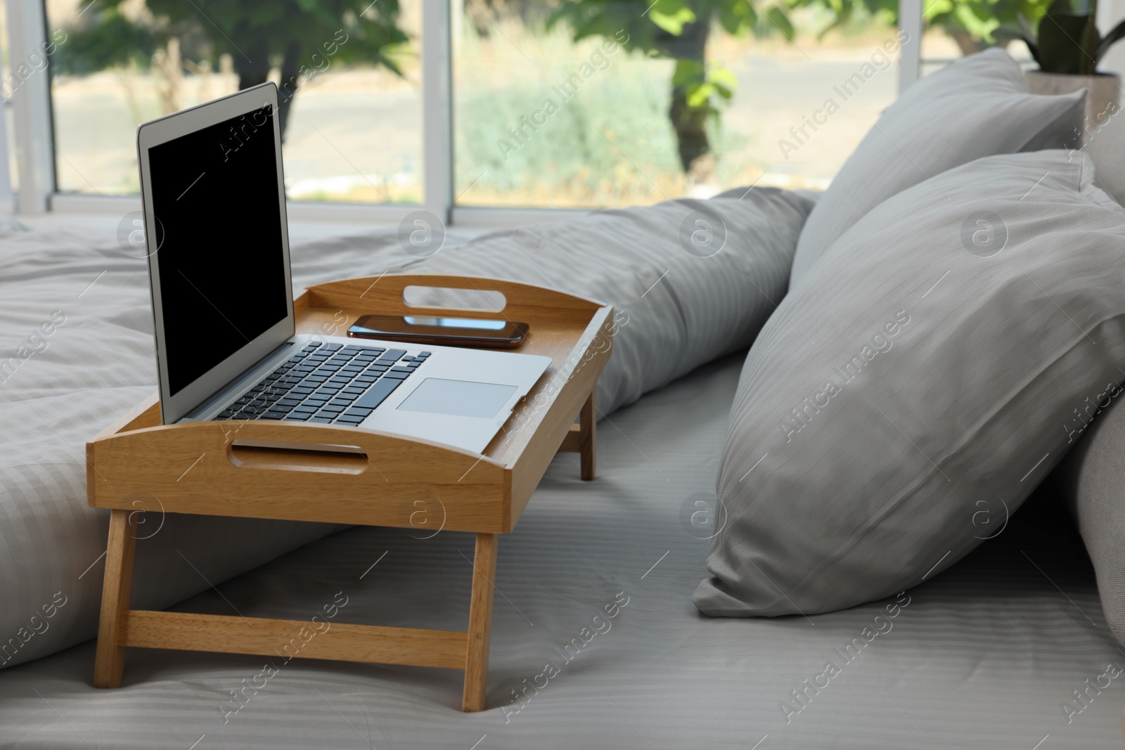 Photo of Wooden tray table with laptop and smartphone on bed indoors