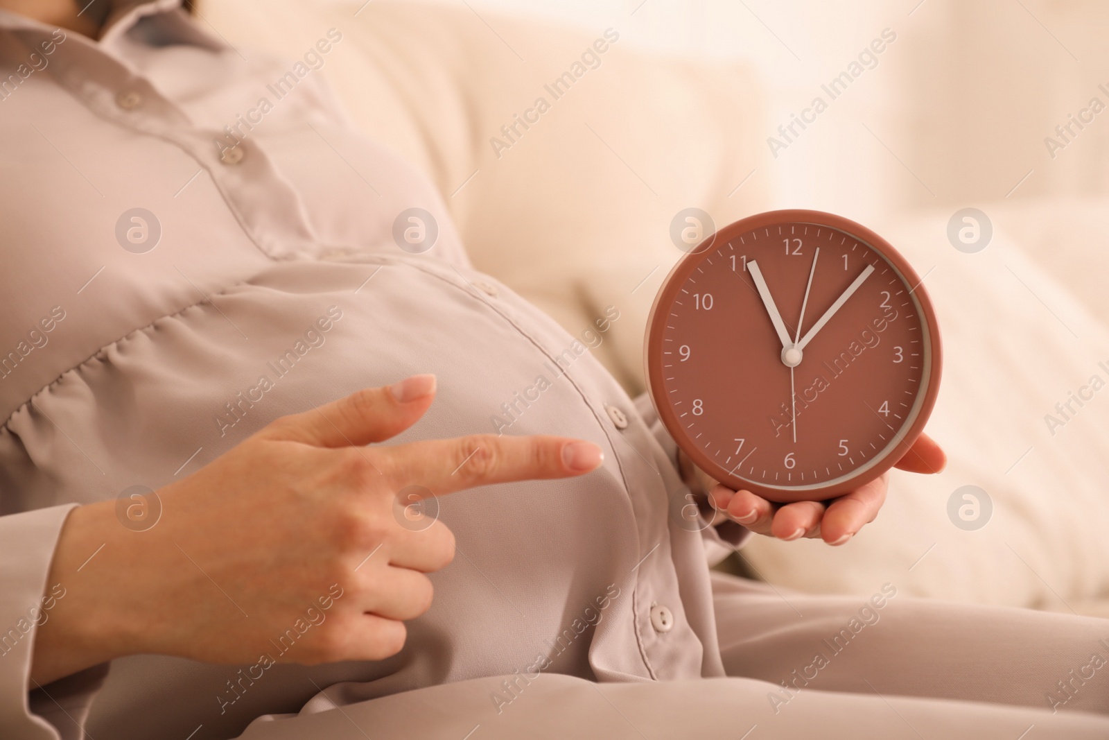 Photo of Young pregnant woman pointing at clock near her belly indoors, closeup. Time to give birth