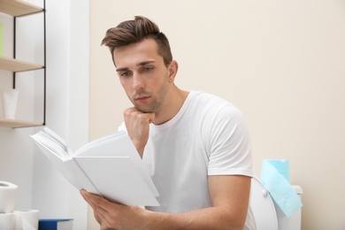 Young man with book sitting on toilet bowl in bathroom