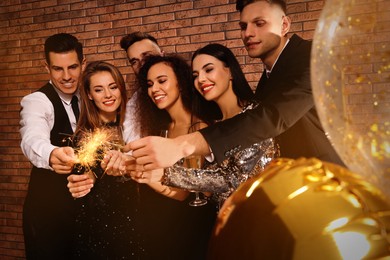 Photo of Happy friends with glasses of sparkling wine and sparklers celebrating New Year indoors