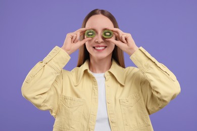 Photo of Young woman holding halves of kiwi near her eyes on purple background