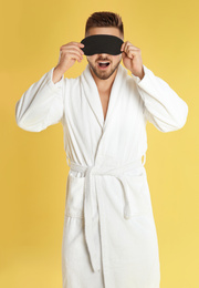 Photo of Young man in bathrobe and eye sleeping mask on yellow background