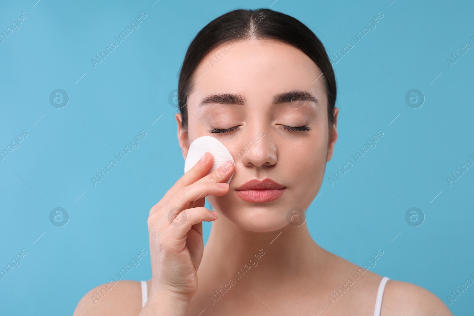 Photo of Beautiful woman removing makeup with cotton pad on light blue background