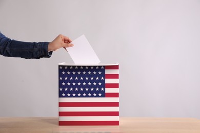 Woman putting her vote into ballot box decorated with flag of USA against light background, closeup