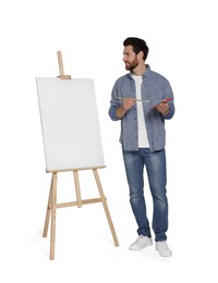 Photo of Happy man with brush and artist`s palette near easel with canvas against white background. Creative hobby