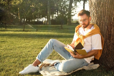 Young man reading book on green grass near tree in park