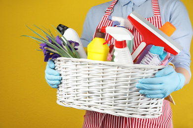 Photo of Woman holding basket with spring flowers and cleaning supplies on yellow background, closeup