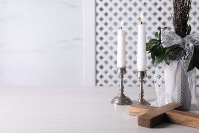 Burning candles, bouquet with willow branches and cross on white wooden table, space for text