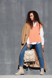 Full length portrait of beautiful African-American woman with stylish beige backpack near grey wall outdoors