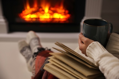 Woman with cup of hot drink and book resting near fireplace at home, closeup