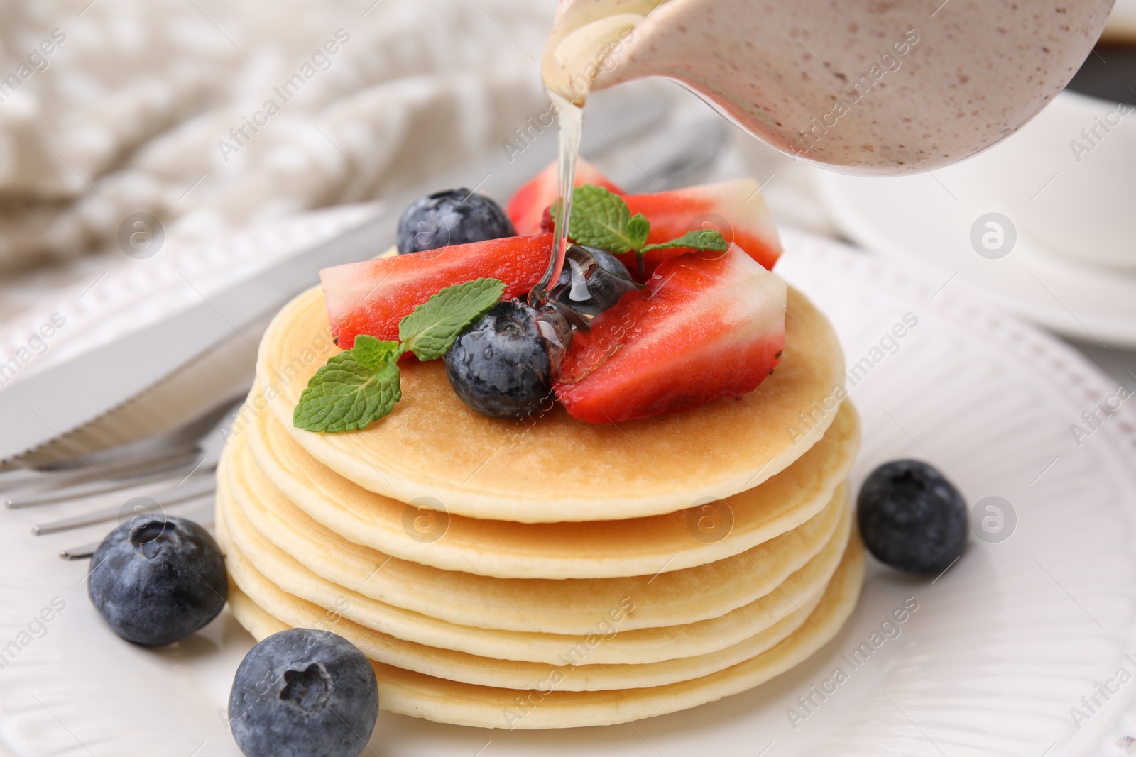 Photo of Pouring honey from jug onto delicious pancakes with strawberries, blueberries and mint at table, closeup