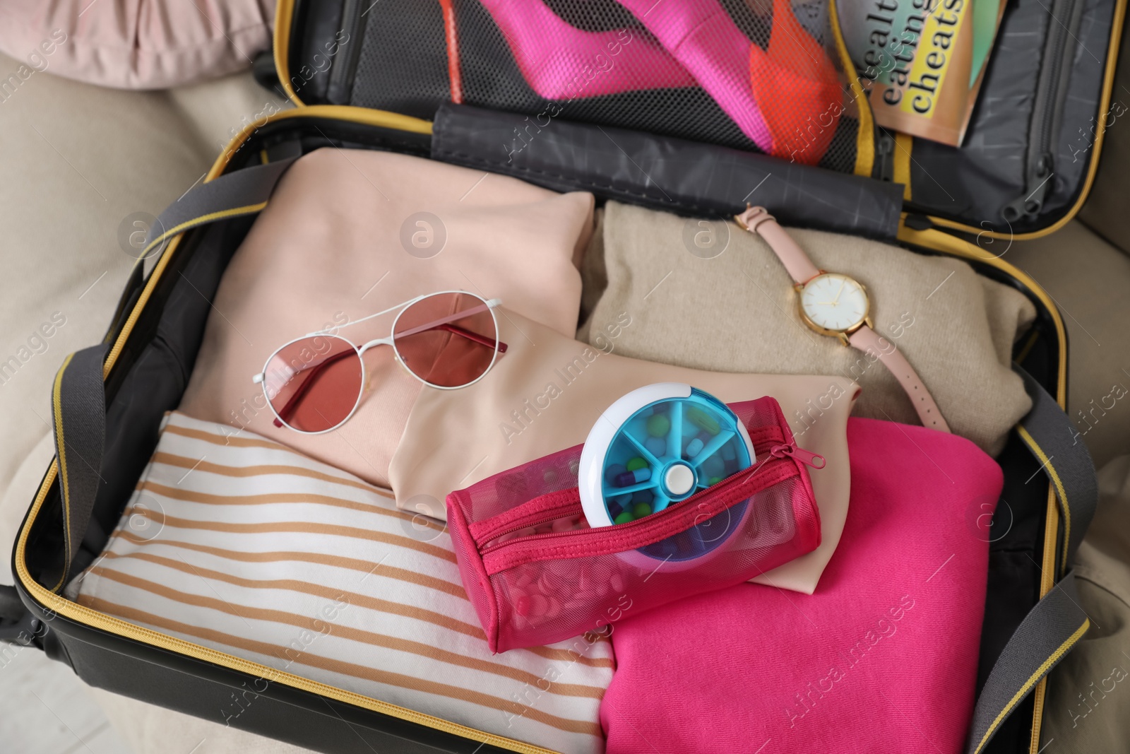 Photo of Open suitcase with packed clothes, accessories and pill box on sofa