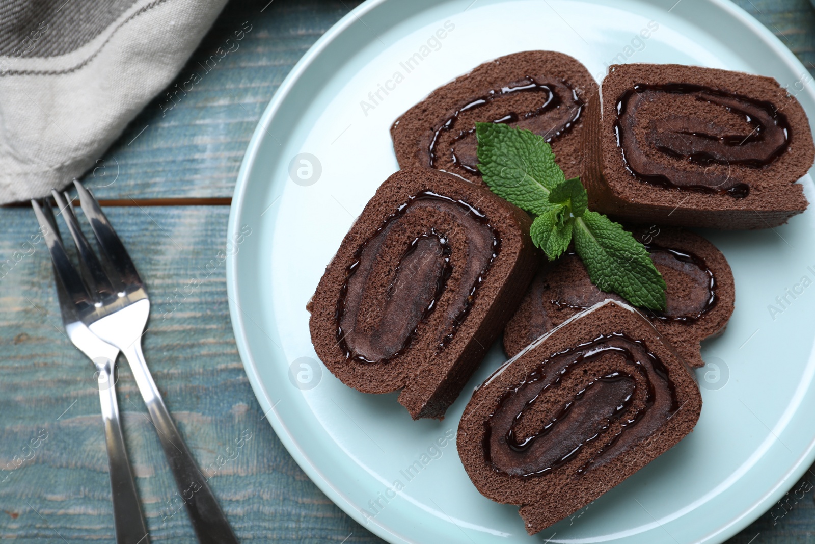 Photo of Tasty chocolate cake roll with cream and mint on blue wooden table, flat lay