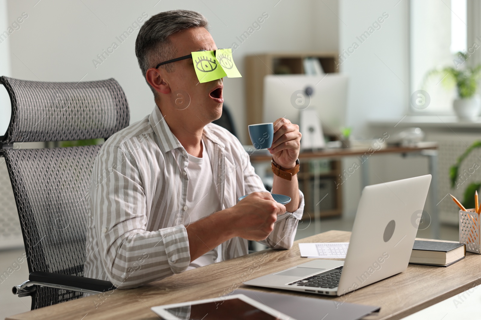 Photo of Man with fake eyes painted on sticky notes holding cup of drink and yawning at workplace in office