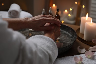 Photo of Woman soaking her hands in bowl of water and flower petals at table, closeup. Spa treatment
