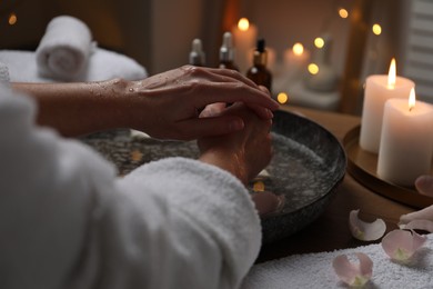 Woman soaking her hands in bowl of water and flower petals at table, closeup. Spa treatment