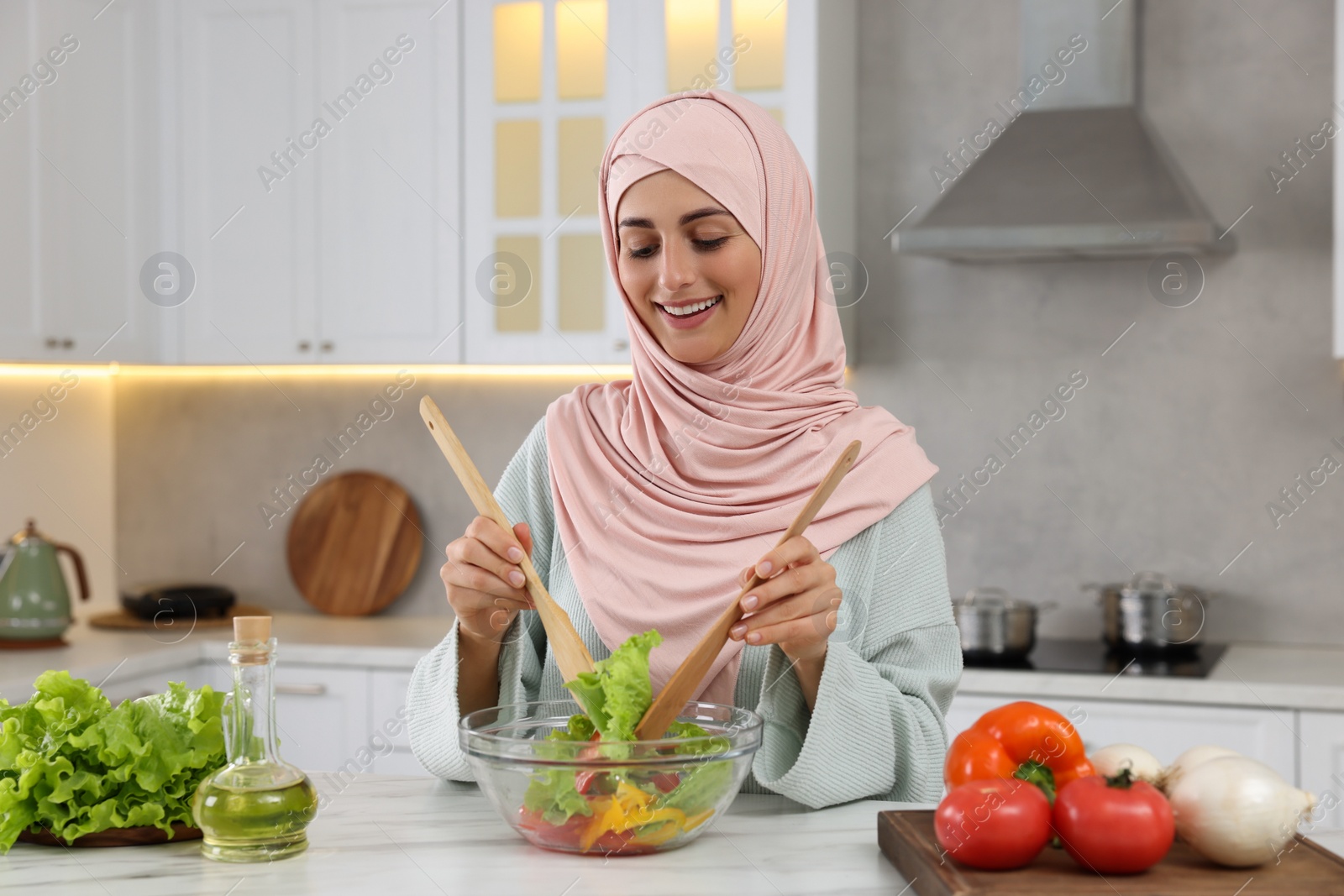 Photo of Muslim woman making delicious salad with vegetables at white table in kitchen