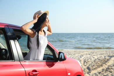 Happy young woman leaning out of car window on beach, space for text. Summer trip