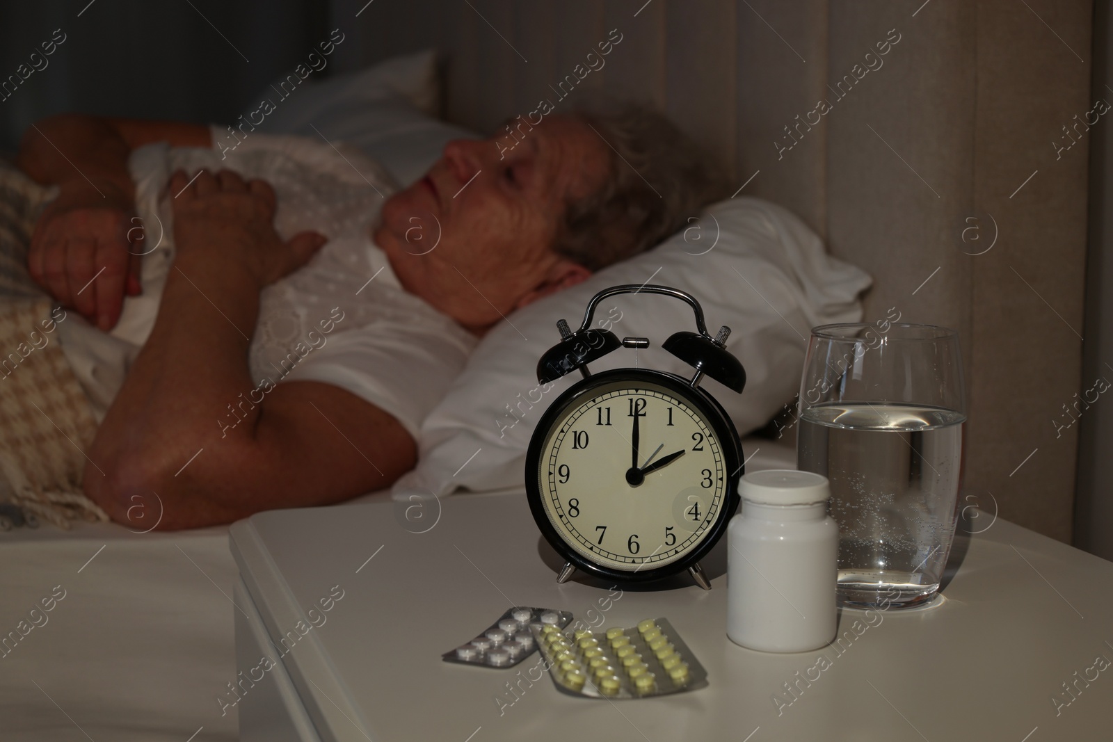 Photo of Elderly woman suffering from insomnia in bed at home, focus on pills and alarm clock