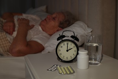 Elderly woman suffering from insomnia in bed at home, focus on pills and alarm clock