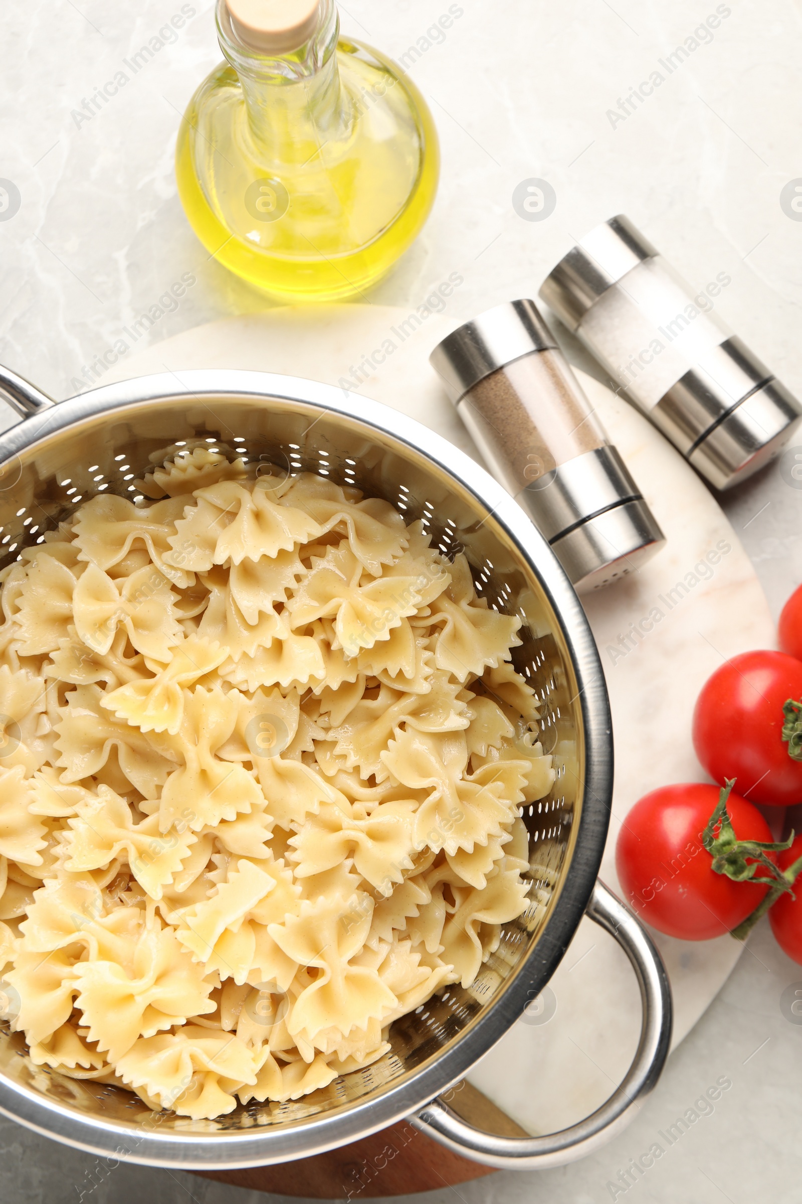 Photo of Cooked pasta in metal colander, oil, spices and tomatoes on light table, flat lay