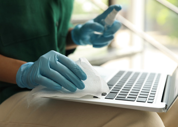 Photo of Woman in latex gloves cleaning laptop with wet wipe indoors, closeup