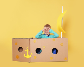Photo of Little child playing with ship made of cardboard box on yellow background