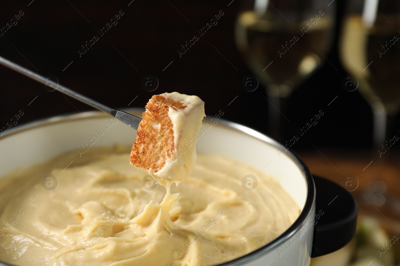 Photo of Dipping piece of bread into fondue pot with melted cheese at table, closeup