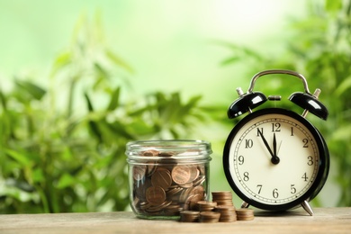Photo of Black alarm clock, glass jar and coins on wooden table. Money savings