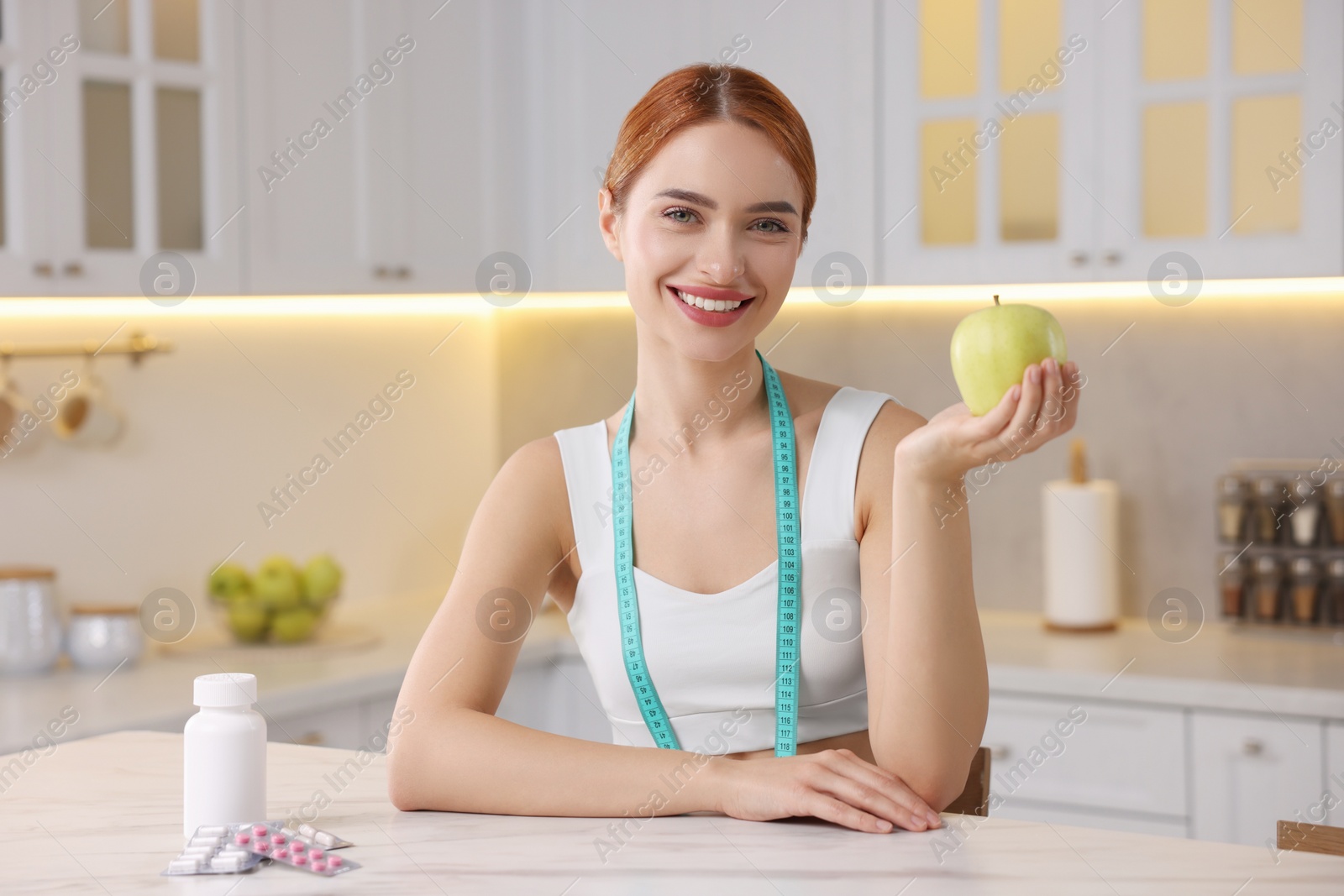 Photo of Young woman with measuring tape and apple at table with pills in kitchen. Weight loss