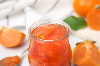 Jar of tasty persimmon jam on white table, closeup