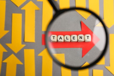 Photo of Recruitment process, searching for employee. View through magnifying glass of red paper arrow and word Talent made of wooden cubes among yellow ones on grey background, flat lay