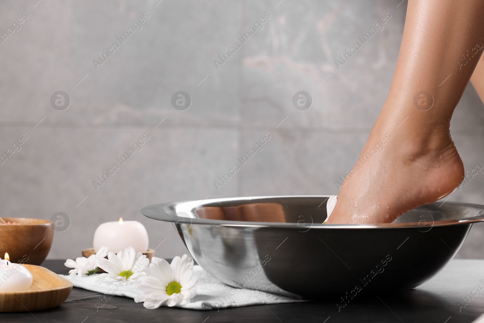 Photo of Woman soaking her foot in bowl with water on dark surface, closeup. Pedicure procedure