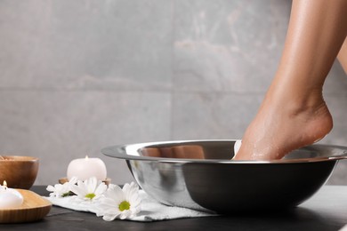 Woman soaking her foot in bowl with water on dark surface, closeup. Pedicure procedure