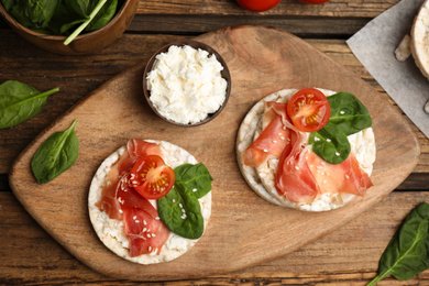 Photo of Puffed rice cakes with prosciutto, tomato and basil on wooden table, flat lay