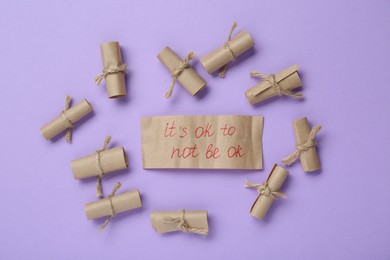Photo of Card with phrase It`s Ok to Not Be Ok and rolled pieces of paper on violet background, flat lay
