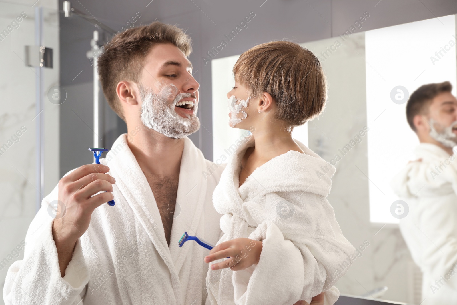 Photo of Dad and son with shaving foam on faces holding razors in bathroom