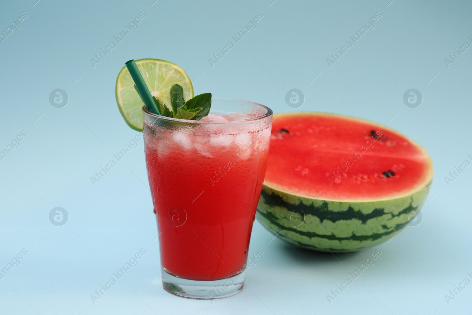 Photo of Glass of delicious drink with lime, ice cubes and cut fresh watermelon on light blue background