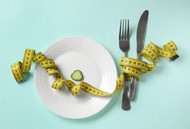 Plate with cucumber slice, cutlery and measuring tape on light blue background, flat lay. Diet concept