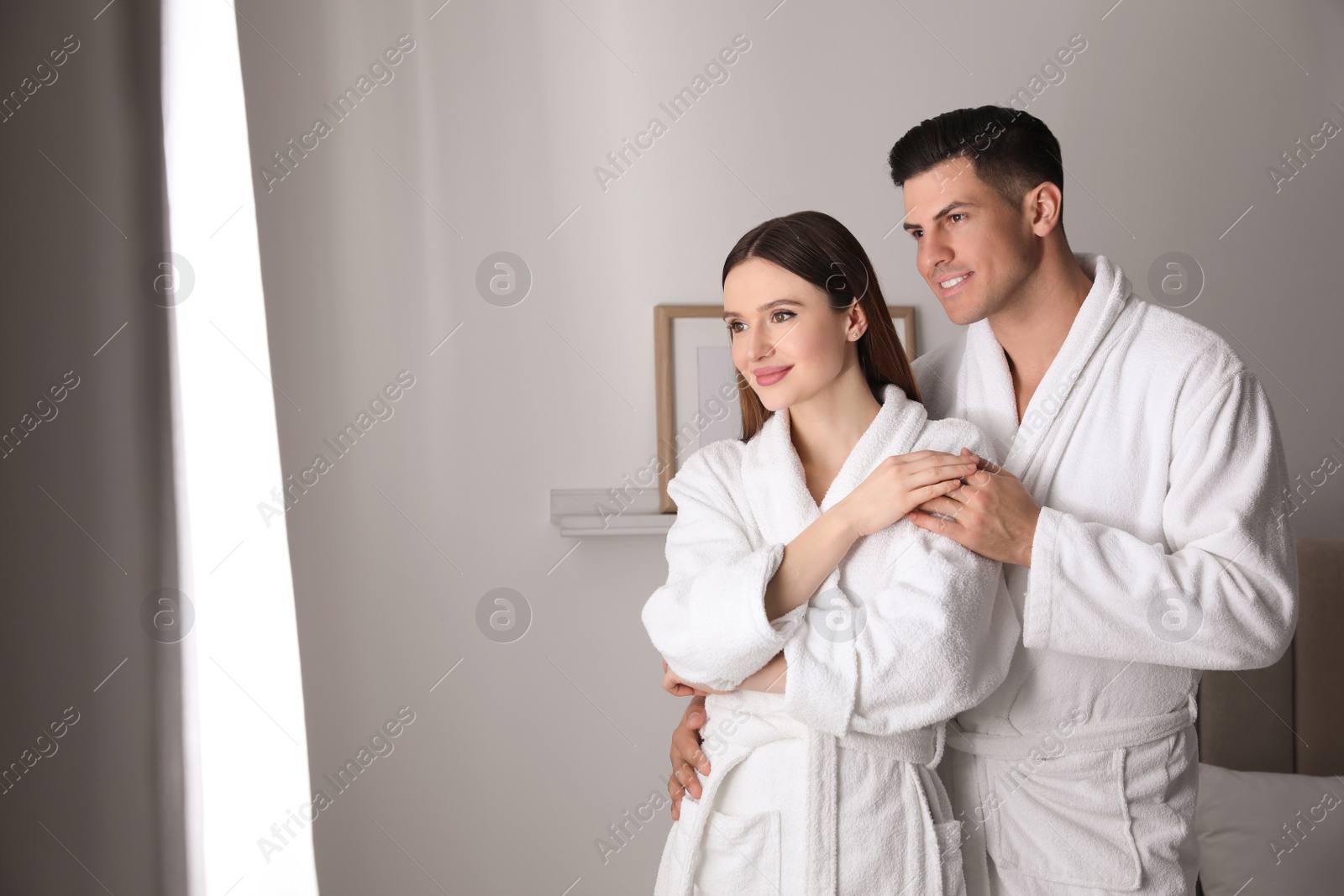 Photo of Happy couple wearing bathrobes near window in room
