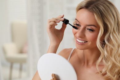 Beautiful makeup. Smiling woman applying mascara in front of mirror indoors